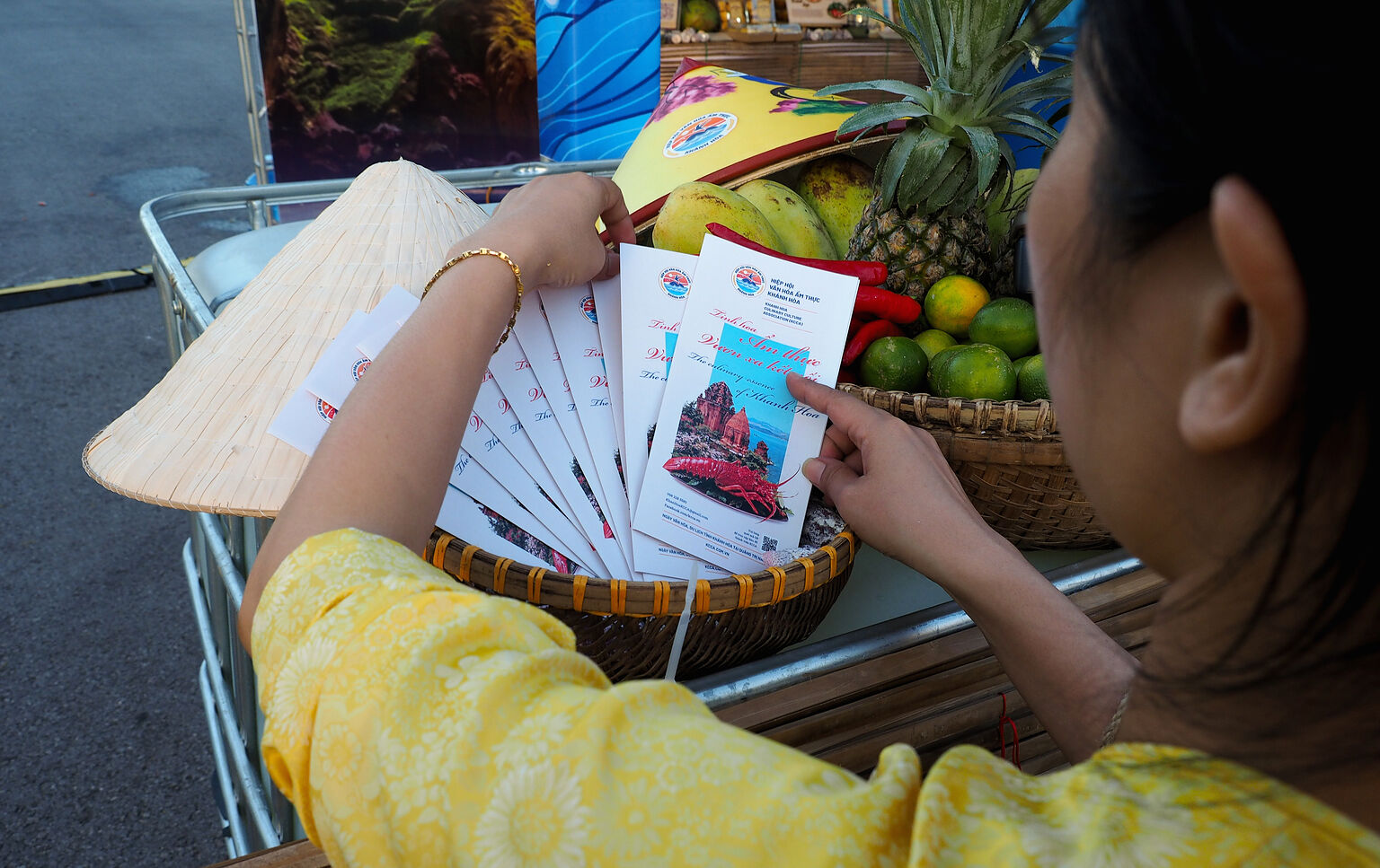 Tay cầm tập brochure của Cẩm nang Văn hóa Ẩm thực Khánh Hòa đặt trên mẹt tre cùng nón lá và trái cây